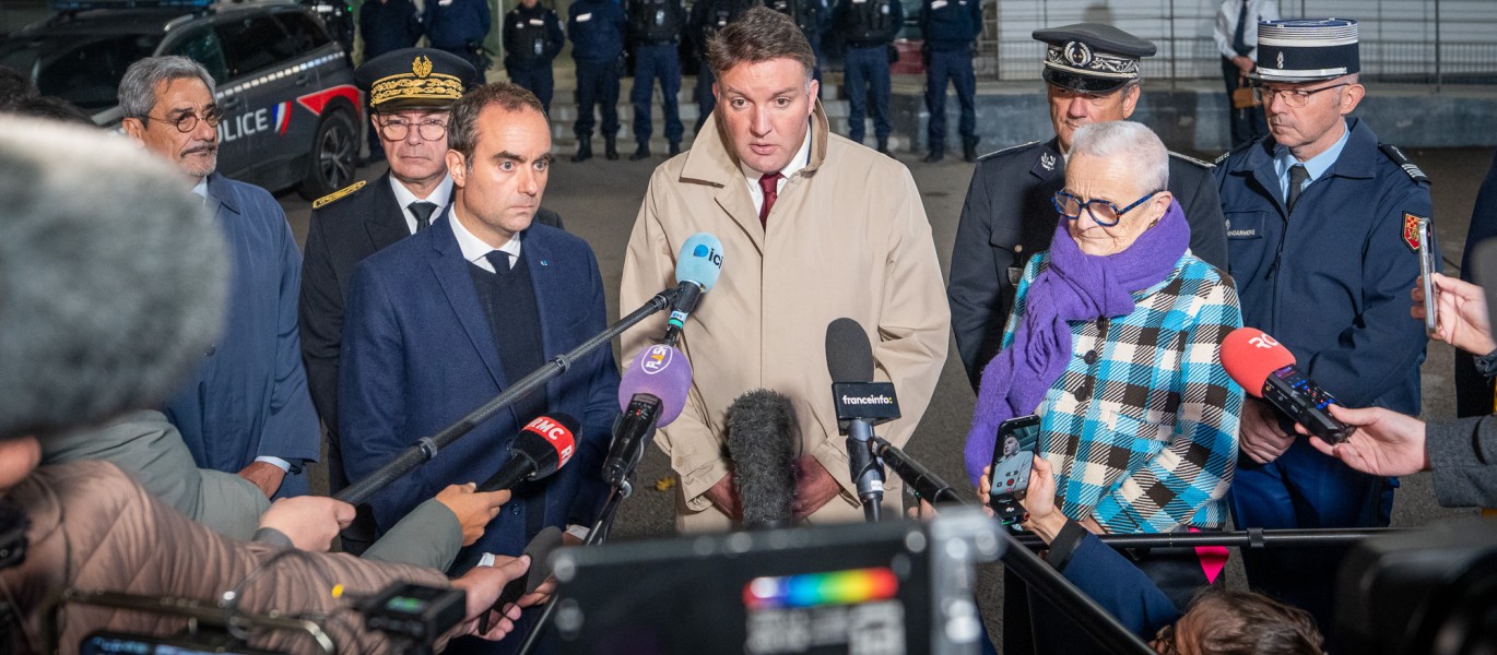 Conférence de presse en extérieur devant un bâtiment sous forte présence policière, avec plusieurs personnes en costume ou uniforme, entourées de nombreux journalistes tendant des micros et caméras. Une femme âgée en manteau à carreaux bleus et écharpe violette se trouve à droite, tandis qu’un homme en manteau beige parle au centre. À l’arrière-plan, une rangée de policiers en uniforme est alignée devant une voiture de police.