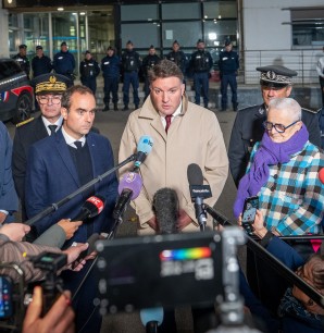 Conférence de presse en extérieur devant un bâtiment sous forte présence policière, avec plusieurs personnes en costume ou uniforme, entourées de nombreux journalistes tendant des micros et caméras. Une femme âgée en manteau à carreaux bleus et écharpe violette se trouve à droite, tandis qu’un homme en manteau beige parle au centre. À l’arrière-plan, une rangée de policiers en uniforme est alignée devant une voiture de police.